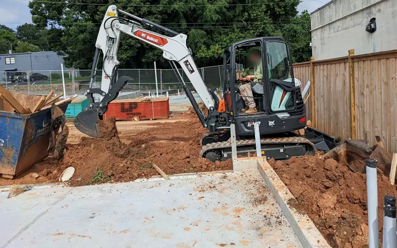 Mini excavator digging and moving red clay next to a new concrete slab on a commercial job