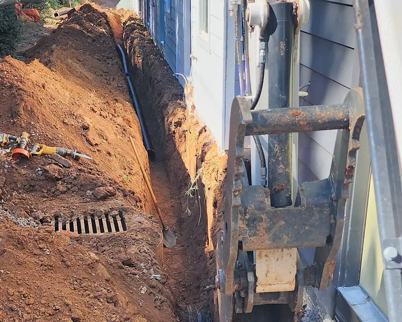 Drainage trench dug along a building foundation with a black drain pipe and a catch basin grate